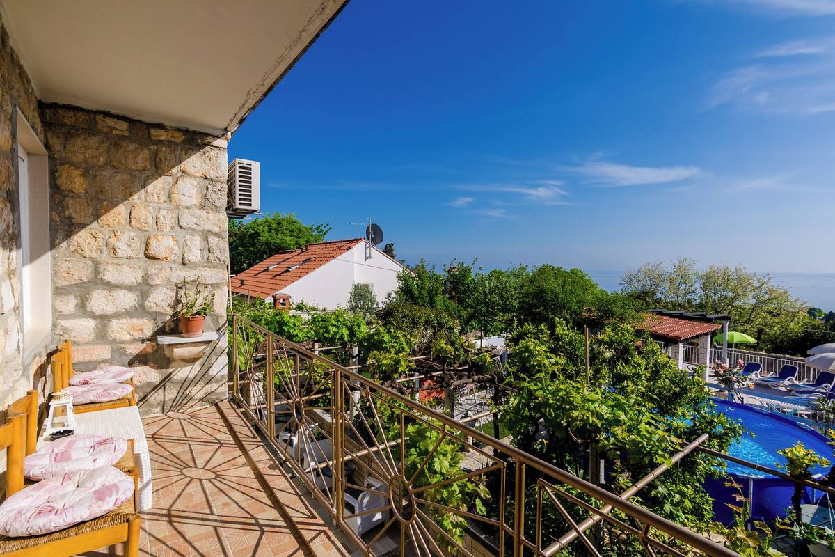 A balcony with a stone wall, a white chair, a small table, and a pool area with lounge chairs and umbrellas.