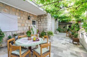 A stone-walled patio with a round table and chairs, surrounded by potted plants and a pergola with vines.