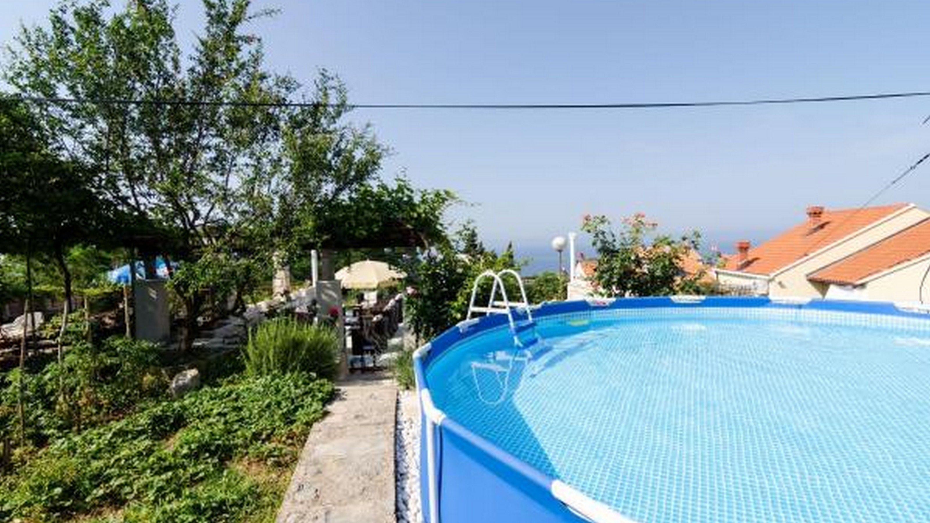 An outdoor swimming pool with a ladder, surrounded by greenery and buildings in the background.