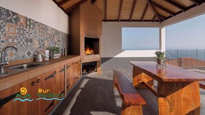 A modern kitchen with a tiled backsplash, a brick fireplace, and a wooden dining table with benches. - Casa da Rosalina (Calheta)
