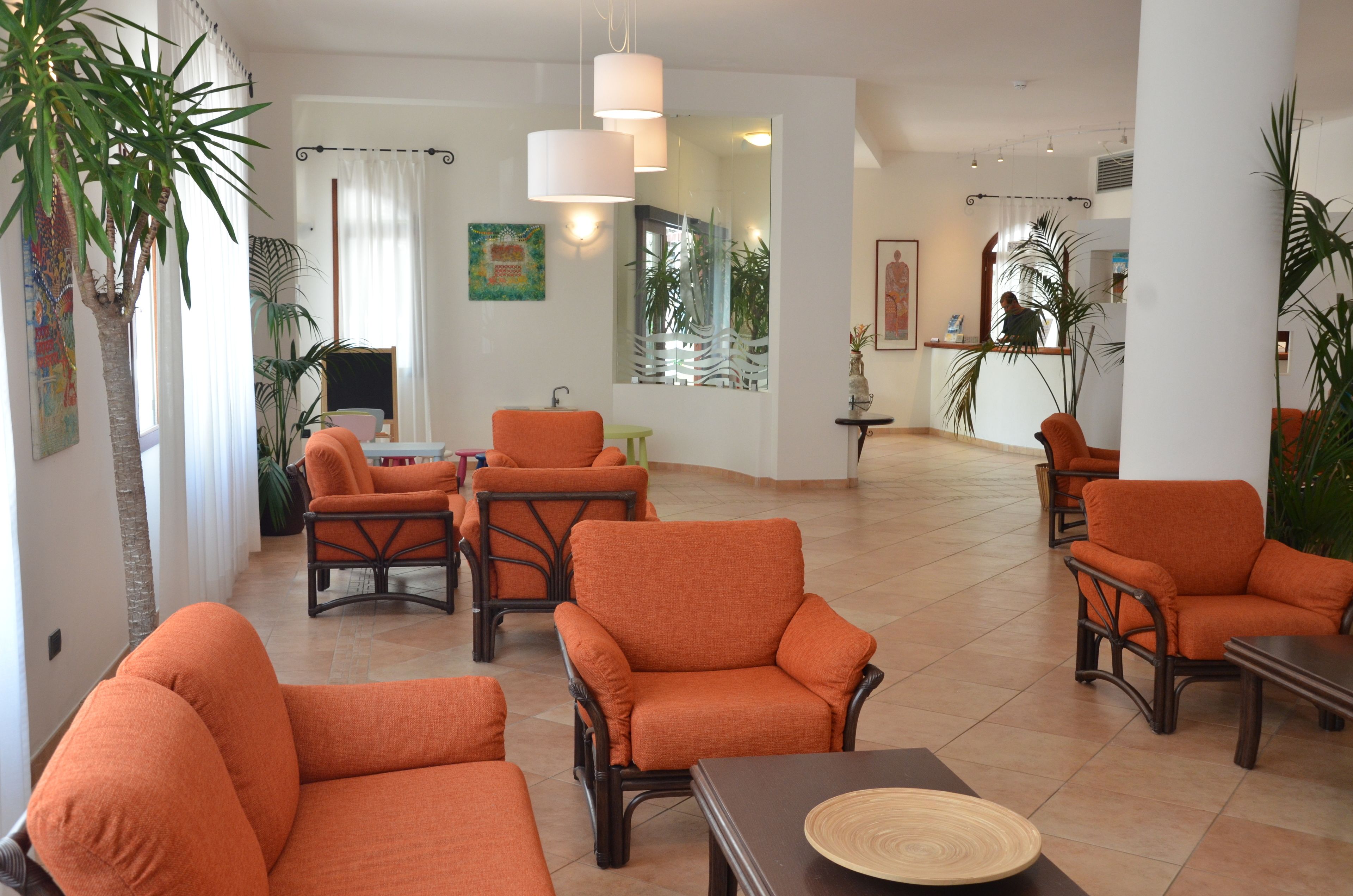 A spacious lobby with orange armchairs and a central table, large windows with curtains, and several potted plants.