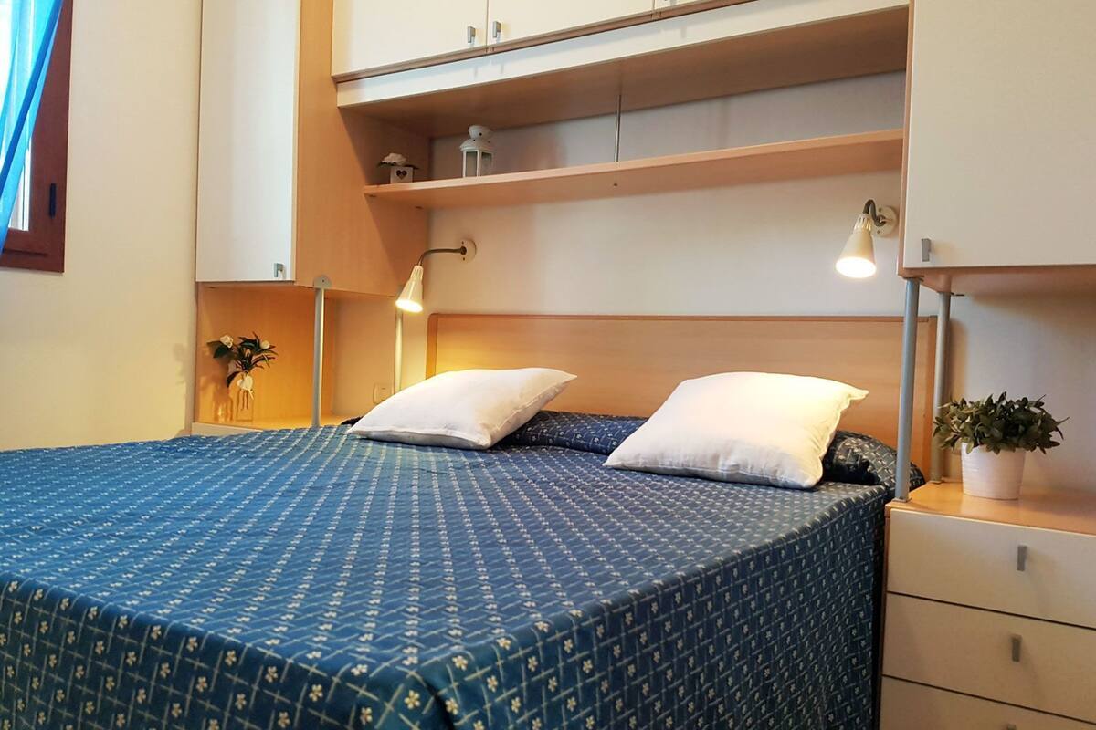 A neatly arranged bedroom with a bed, bedside lamps, a nightstand, and a shelf with decorative items.