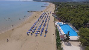 Vista aérea de una playa con hileras de sombrillas azules y tumbonas, una piscina y una zona boscosa al fondo.