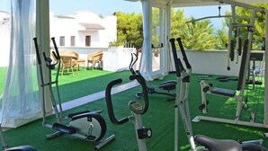 Gimnasio al aire libre con equipamiento de ejercicio, un área techada y vistas a un edificio blanco y árboles.