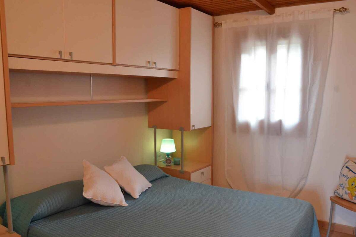 A bedroom with a bed, two pillows, a nightstand with a lamp, a small table with a picture frame, and a window with a sheer curtain.
