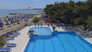 Una piscina con carriles azules, rodeada de sillas de descanso y sombrillas, con una playa y árboles al fondo.