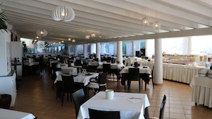 Un restaurante espacioso con numerosas mesas y sillas, ventanas amplias y lámparas colgantes.