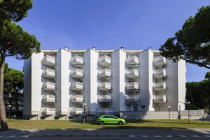 Un immeuble à plusieurs étages doté de balcons, une voiture verte garée devant et des arbres tout autour.