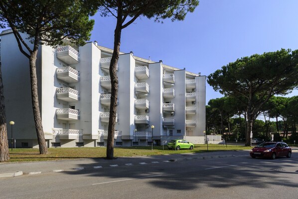 Un immeuble résidentiel à plusieurs étages, doté de balcons, avec une voiture verte garée devant et un ciel bleu dégagé.