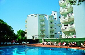 Une piscine avec des chaises longues et un immeuble à plusieurs étages doté de balcons.
