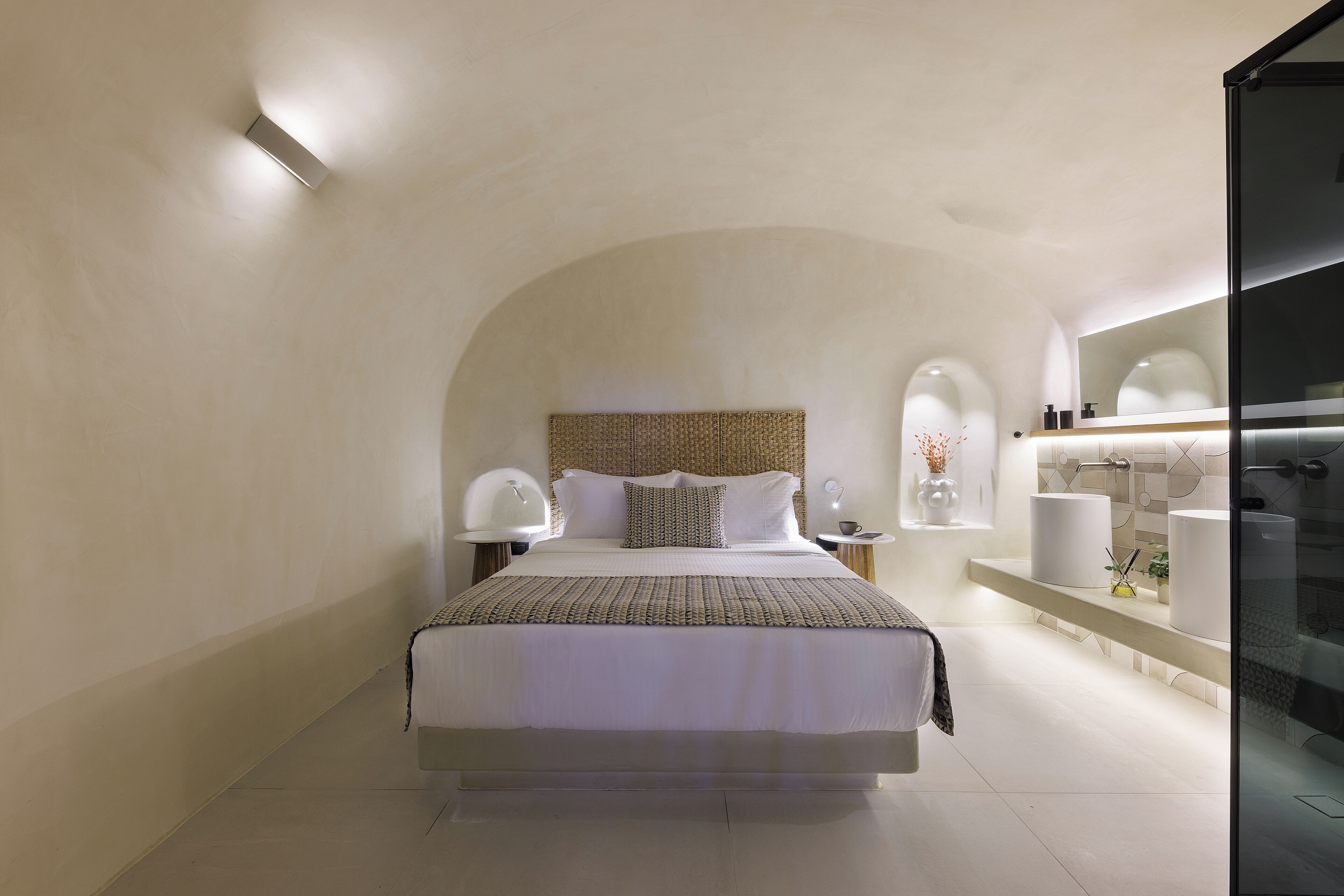 A modern hotel room with a large bed, a bedside table with a lamp, a bathroom with a large mirror and a sink.