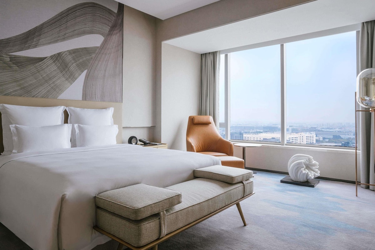 A modern hotel room with a large bed, a seating area, a view of the city, and a decorative sculpture.