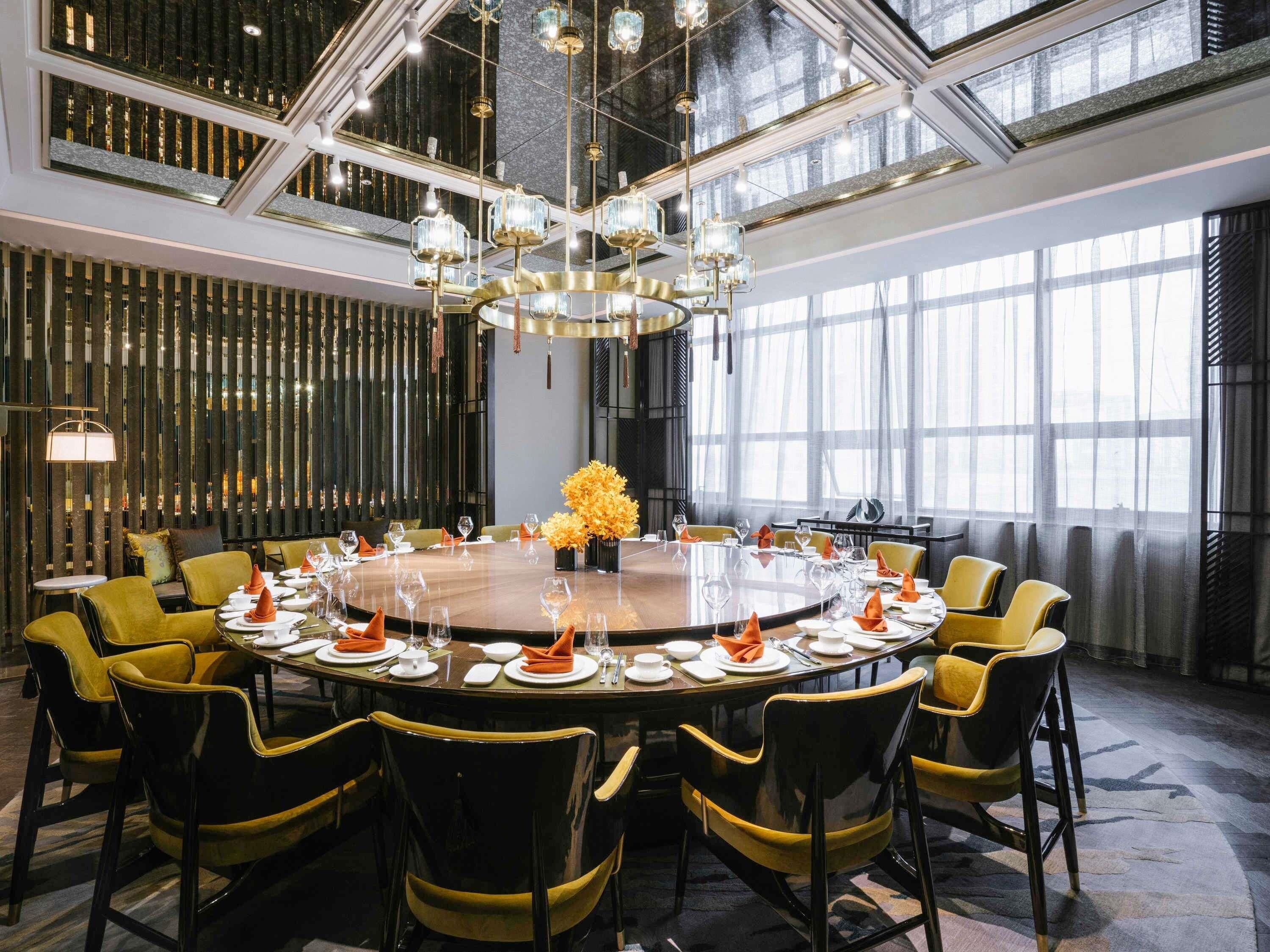 Un comedor amplio con una mesa redonda dispuesta para una comida, rodeado de sillas y una gran lámpara de araña colgando.