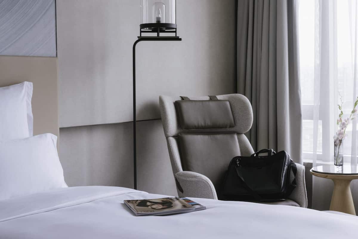 A hotel room with a bed, a chair, a lamp, a handbag, and a table with a vase.