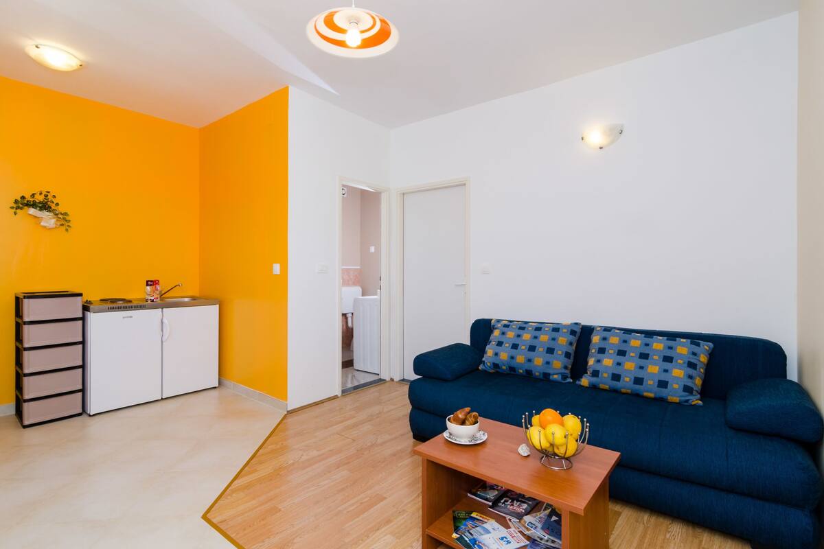 A compact living space with a kitchenette, a blue sofa with patterned cushions, and a wooden coffee table with fruits and magazines.