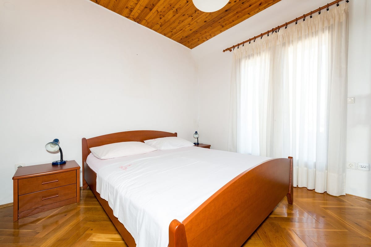 A bedroom with a wooden bed, a nightstand with a lamp, and a window with curtains.