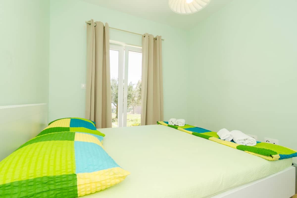 A room with a bed, a window with curtains, and a view of greenery outside.
