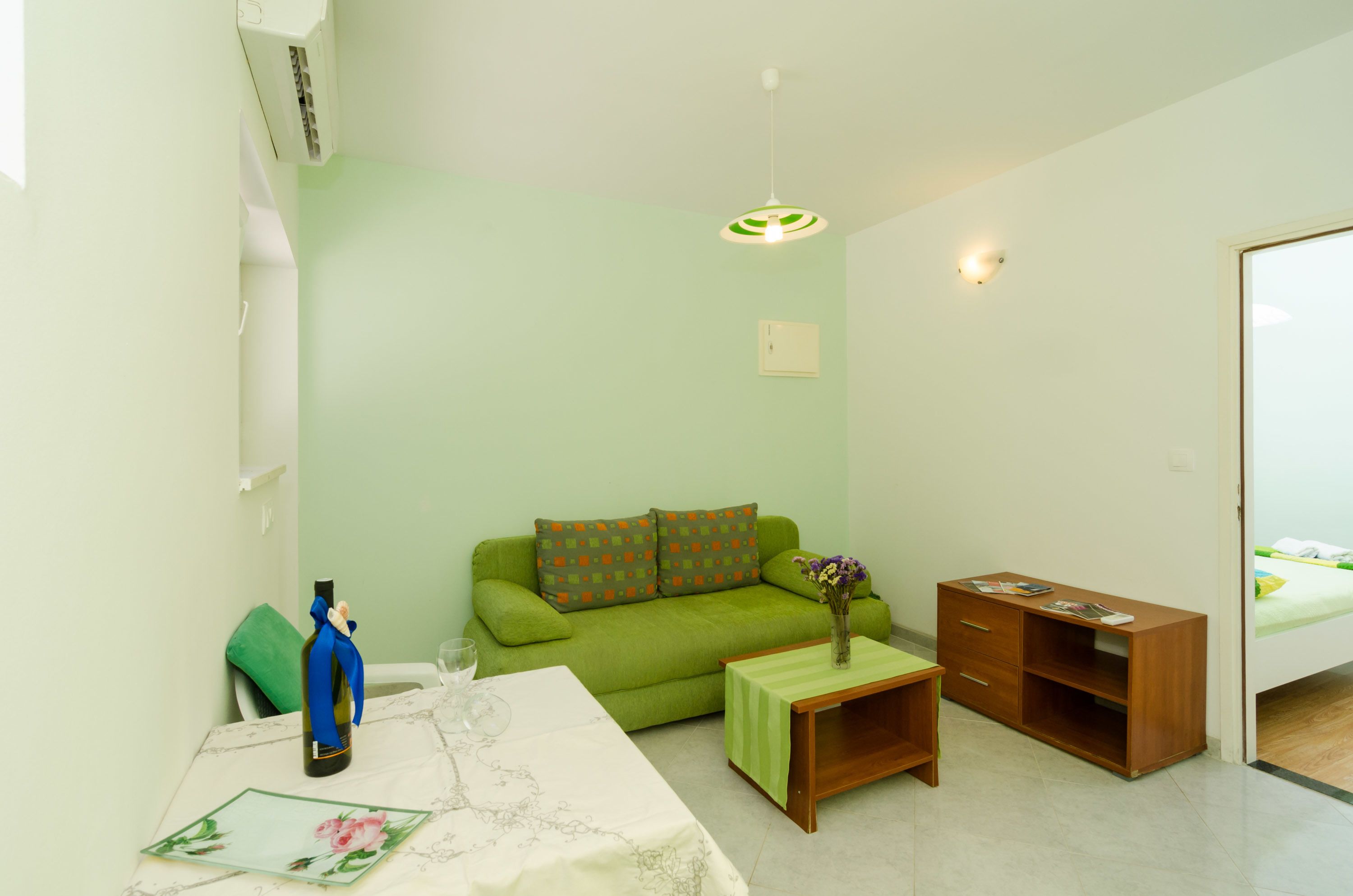 A small, well-lit room with a green sofa, a wooden table with a vase of flowers, and a bed in the background.