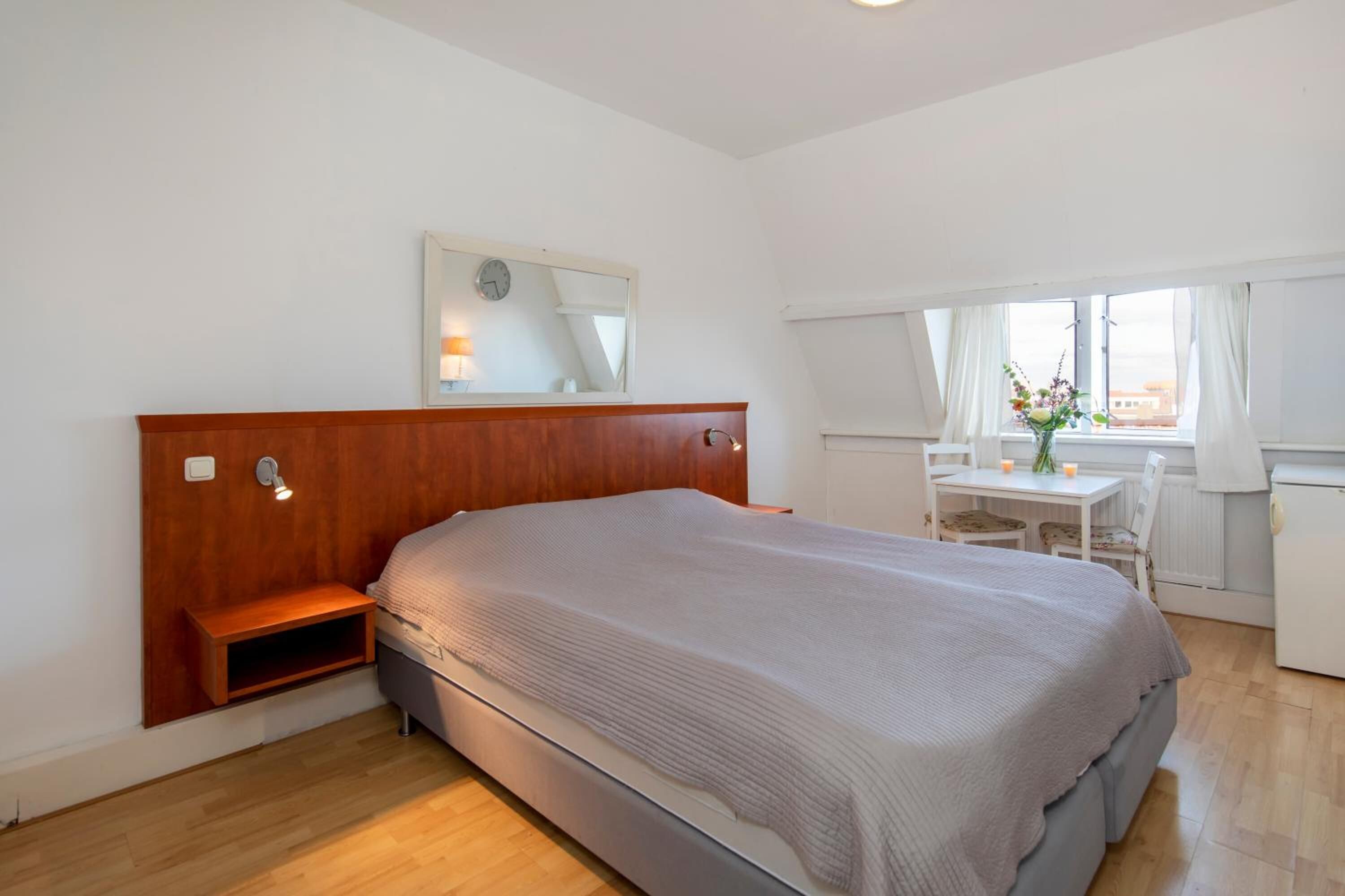 A bedroom with a wooden headboard, a bedside table, a small shelf, a mirror, a window with curtains, and a view of the outside.