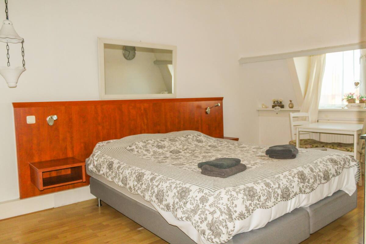 A bedroom with a wooden headboard, a bed with a floral bedspread, a bedside table, a mirror, and a window with curtains.