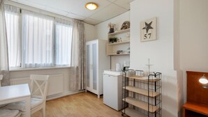 Una habitación pequeña y bien iluminada con una mesa blanca, una silla, un radiador, un electrodoméstico blanco pequeño y un estante metálico con un adorno en forma de estrella y el número 57 en la pared.