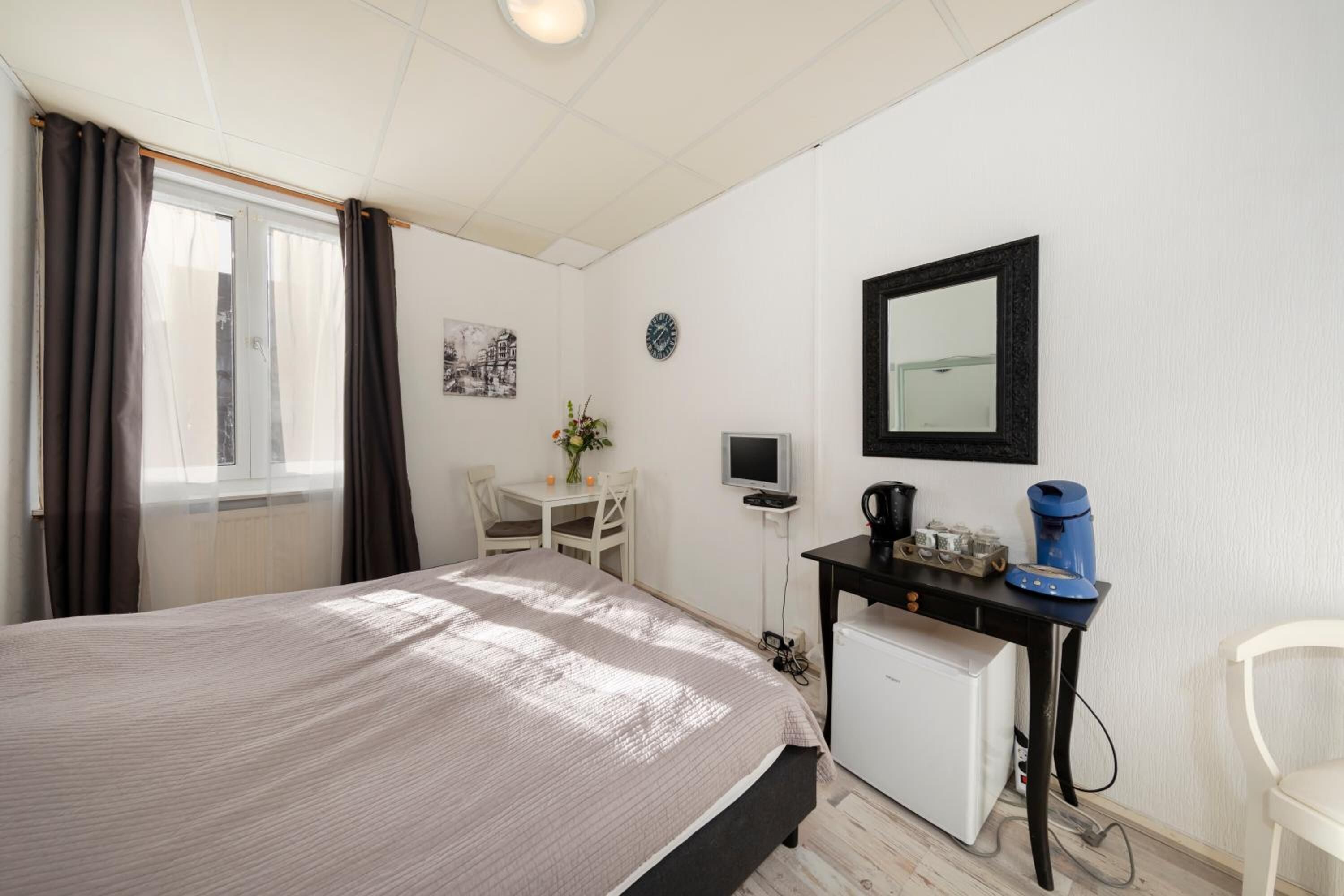 A bedroom with a bed, a desk with a computer, a small fridge, a mirror, and a window with curtains.