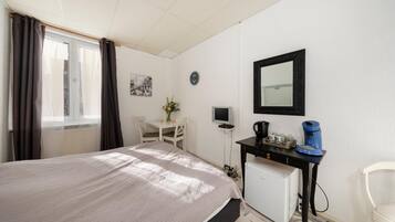 Ein Schlafzimmer mit einem Bett, einem Schreibtisch mit Computer, einer kleinen Kühlbox, einem Spiegel und einem Fenster mit Vorhängen.