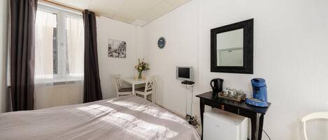 A bedroom with a bed, a desk with a computer, a small fridge, a mirror, and a window with curtains.