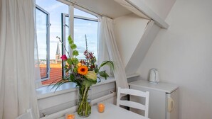Una habitación pequeña y luminosa con una mesa, una silla, un florero y vistas a un edificio a través de una ventana.