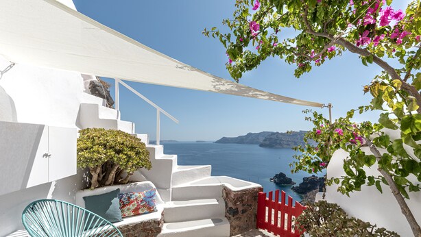 A white staircase leading to a balcony overlooking a body of water, with a red gate and a blue chair. - White Cave Villa by Caldera Houses (Santorini)