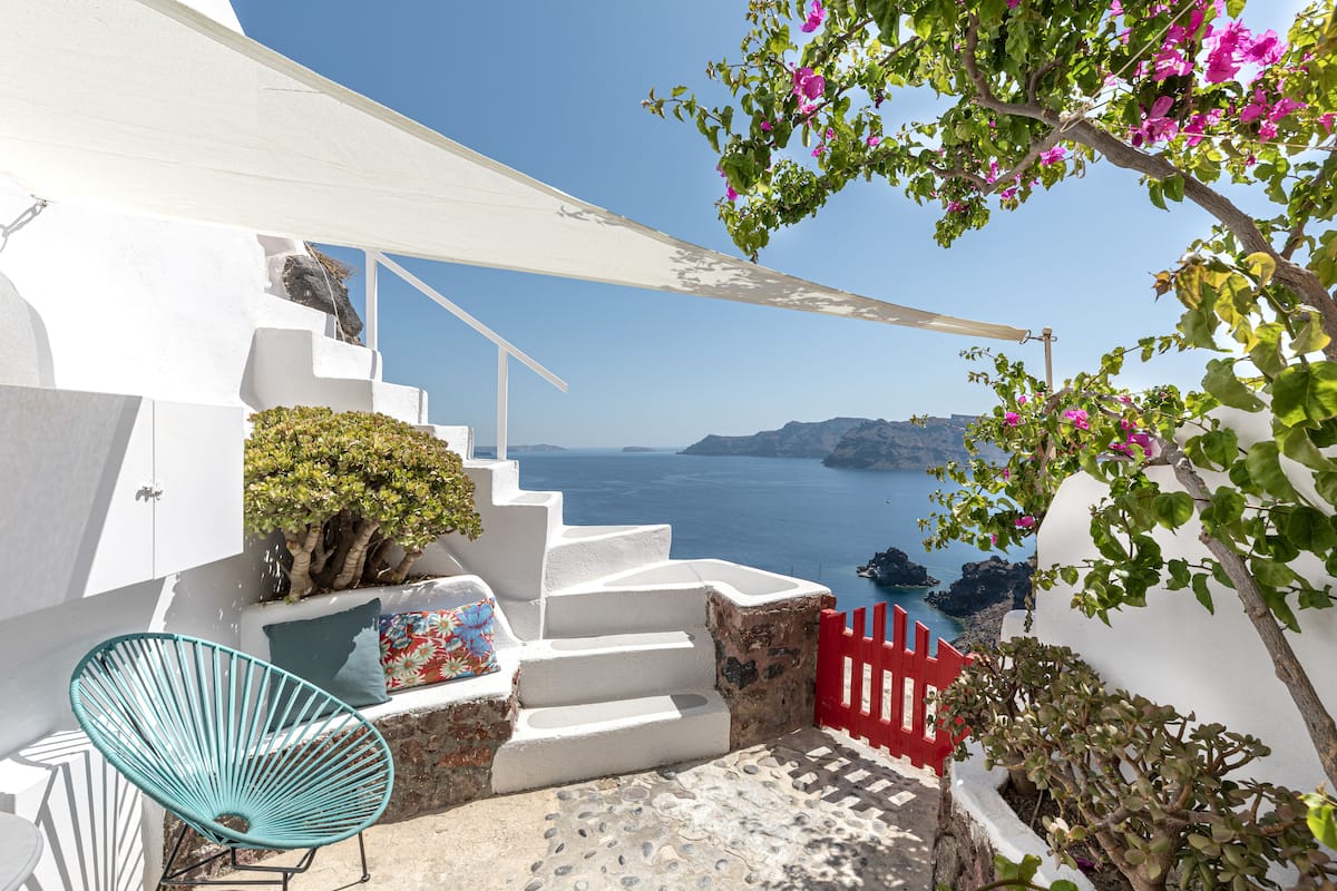 A white staircase leading to a balcony overlooking a body of water, with a red gate and a blue chair.