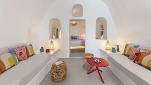 A modern, minimalist living room with white walls, a central archway, and a cozy seating area with a red table and woven ottoman. - White Cave Villa by Caldera Houses (Santorini)