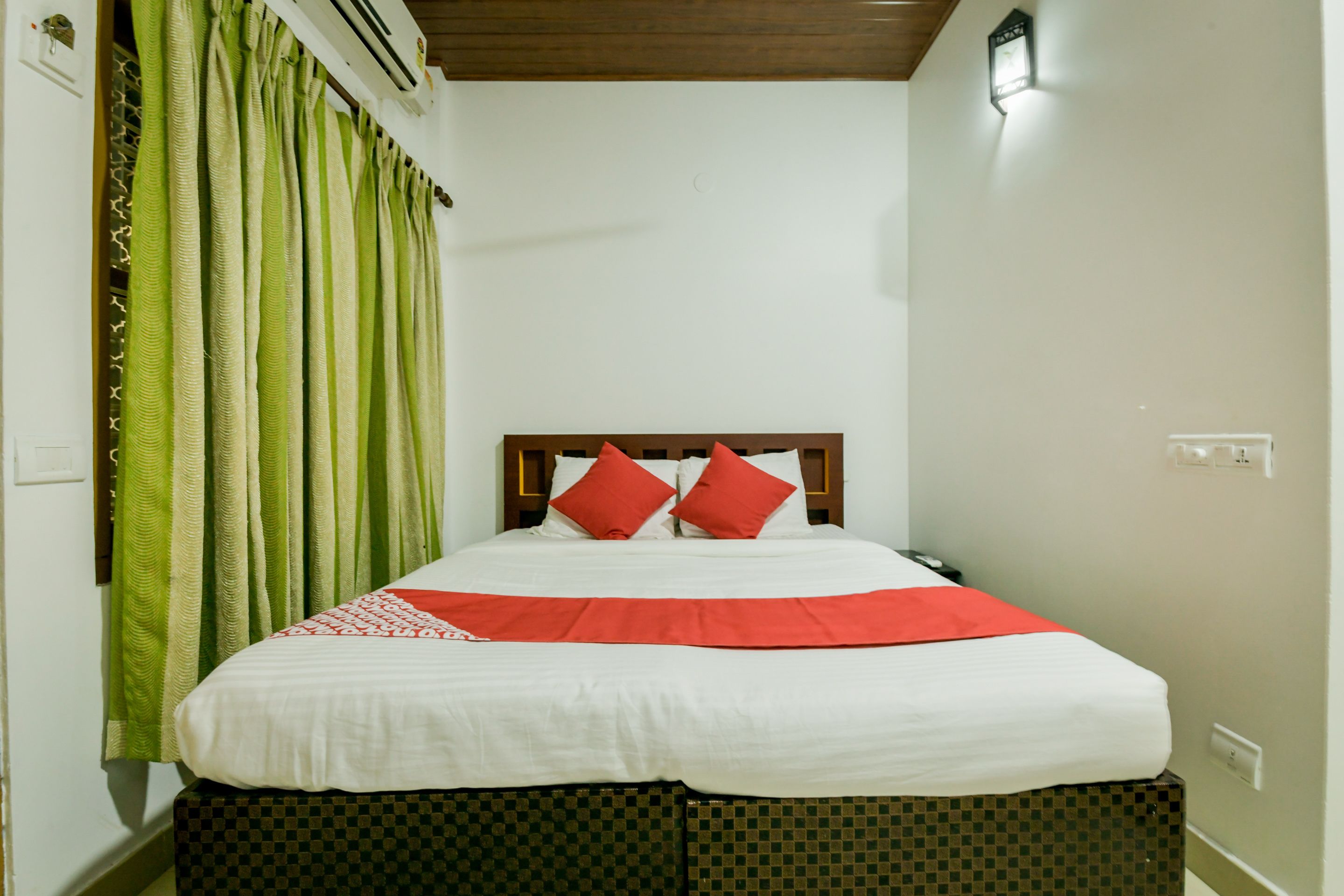 A neatly made bed with a checkered ottoman, green curtains, and a wooden headboard.