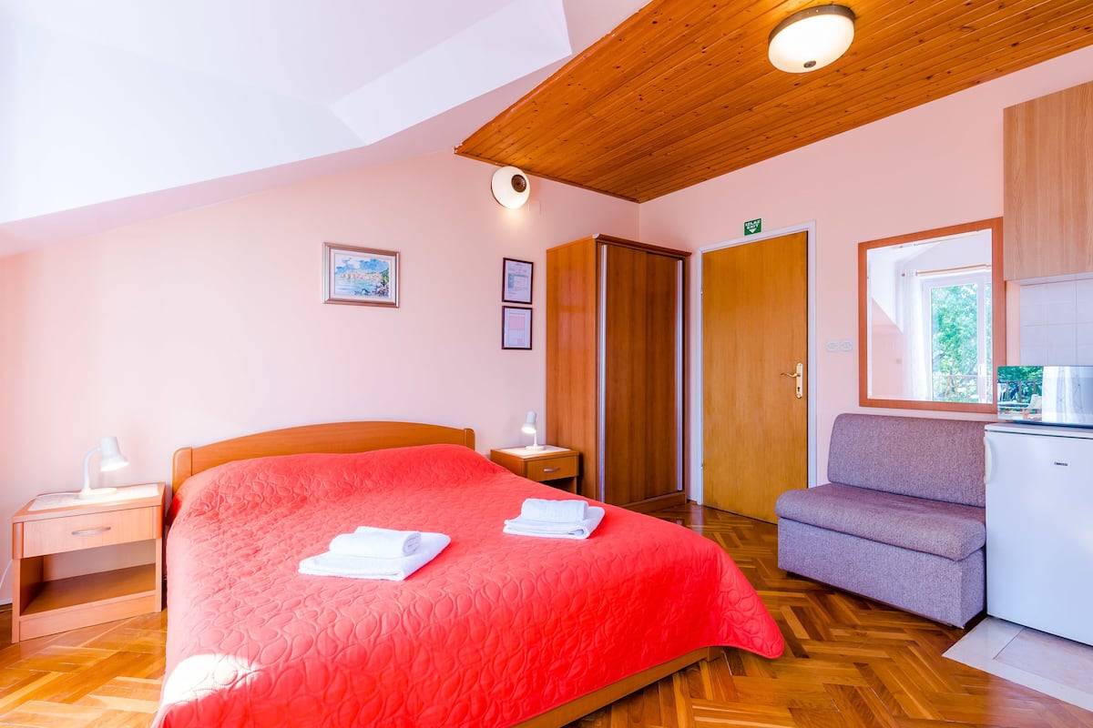 A bedroom with a wooden ceiling, a bed with a red bedspread, a nightstand with a lamp, a sofa, and a small kitchenette.