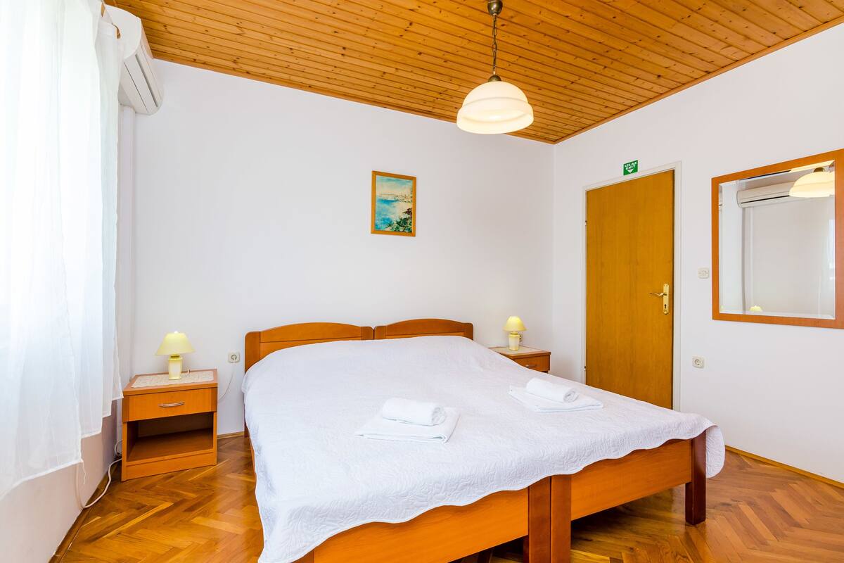 A neatly made bed with a white bedspread and two rolled towels, a wooden nightstand with a lamp, a wooden door, and a framed picture on the wall.
