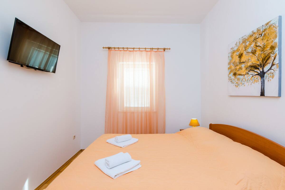 A hotel room with a bed, a television mounted on the wall, a window with sheer curtains, and a painting of a tree.