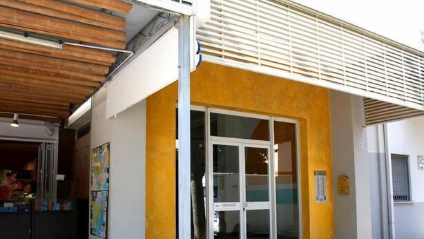 A building with a yellow facade, a large glass door entrance, and a covered area with a wooden ceiling. - Condominio Sabbiadoro (Lignano Sabbiadoro)