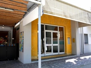 A building with a yellow facade, a large glass door entrance, and a covered area with a wooden ceiling. - Condominio Sabbiadoro (Lignano Sabbiadoro)