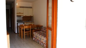 A compact kitchen with wooden cabinets and a dining area with a patterned sofa. - Condominio Sabbiadoro (Lignano Sabbiadoro)