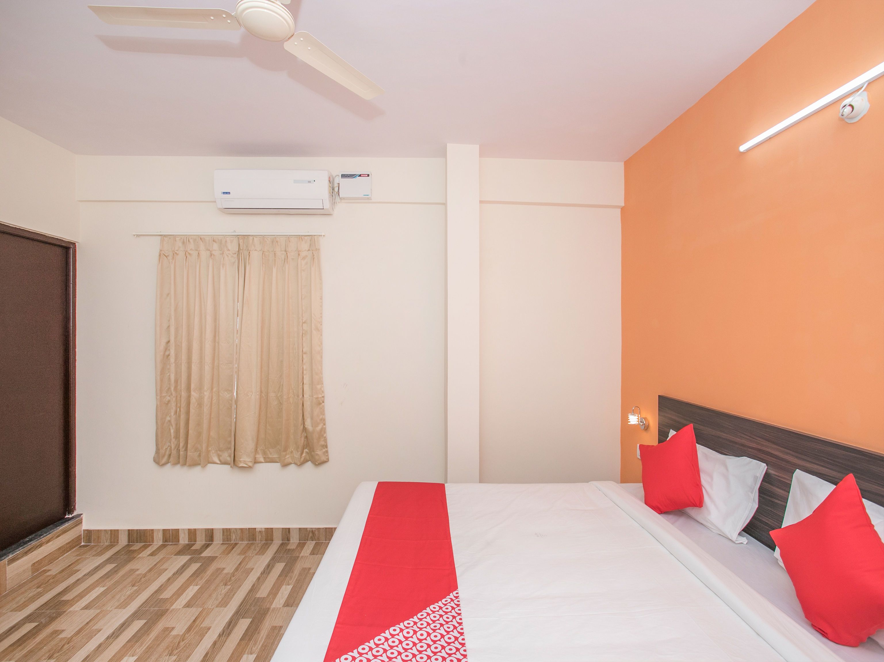 Ein Hotelzimmer mit einem Bett, einem Deckenventilator, einer Klimaanlage, einem Fenster mit Vorhängen und einer wandmontierten Leuchte.