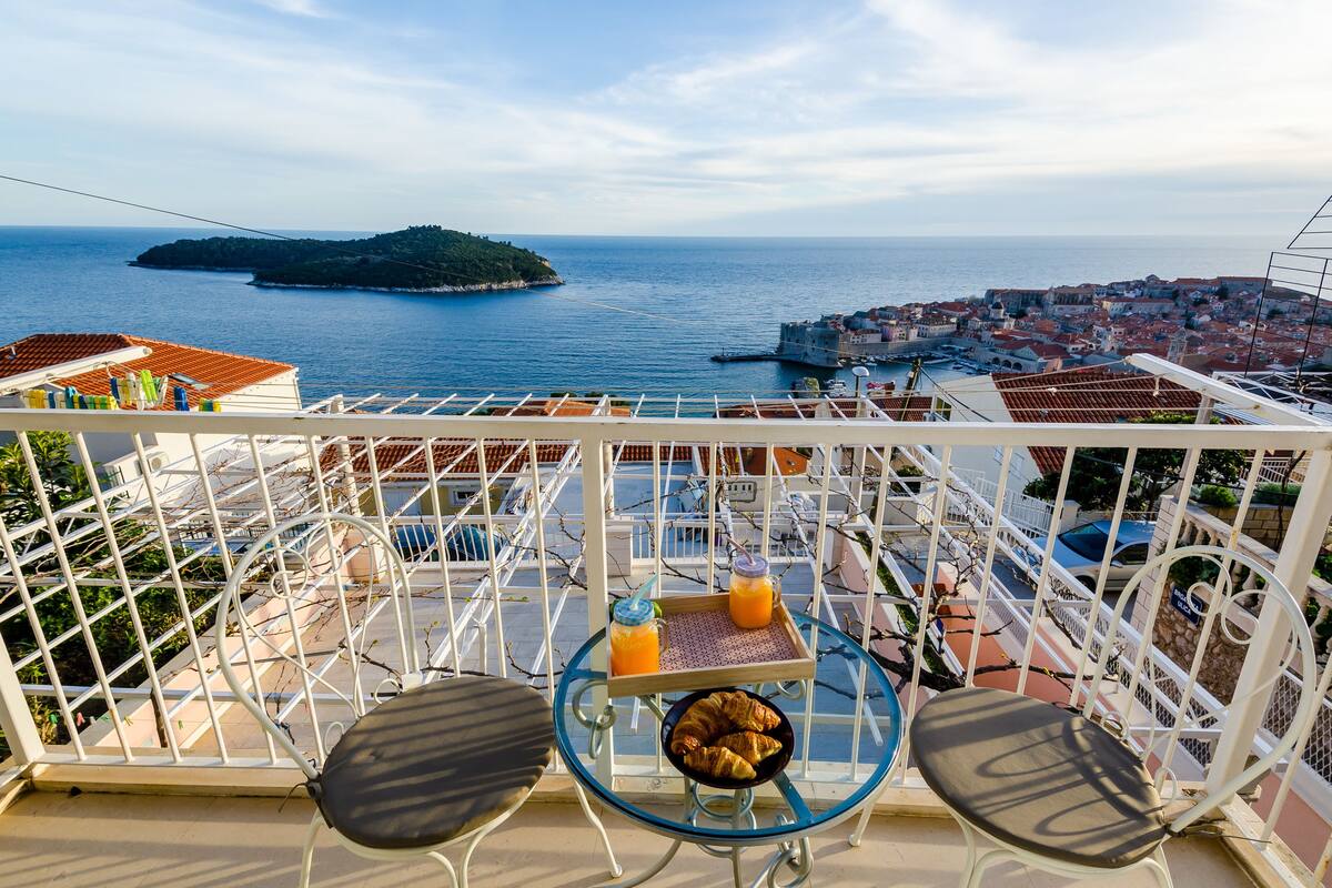 A balcony with a table, two chairs, a tray with croissants and a drink, and a view of the sea and a distant island.