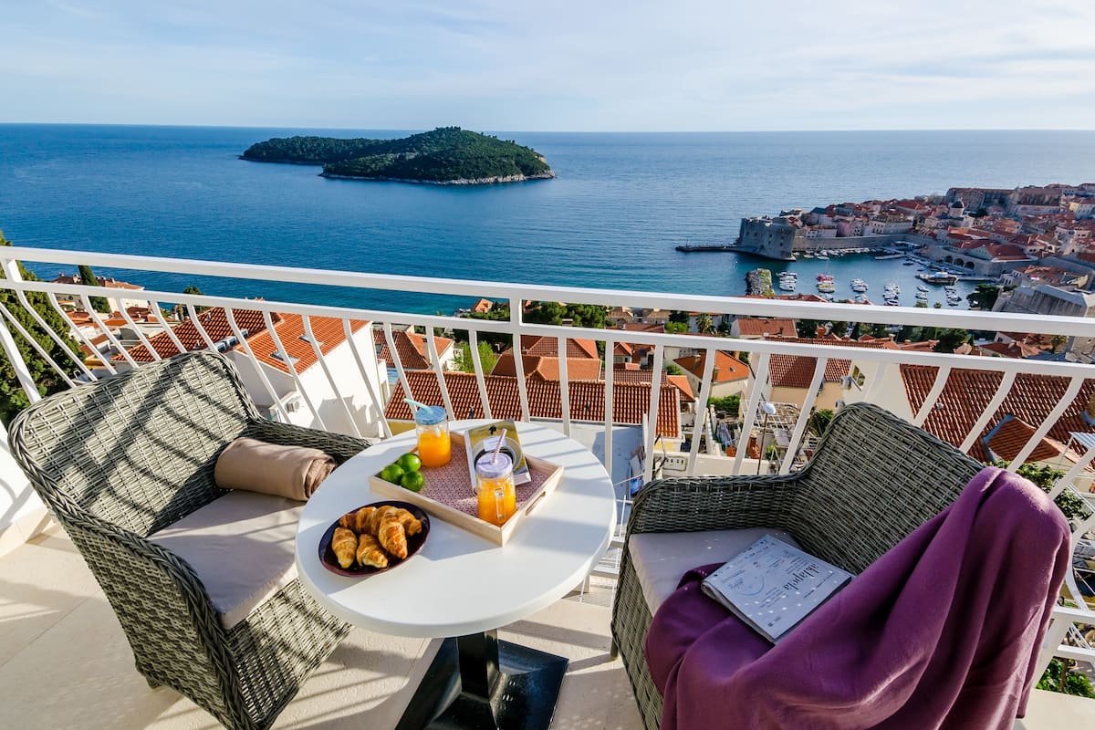 A balcony with wicker furniture, a table with croissants and juice, and a view of the sea and a distant island.