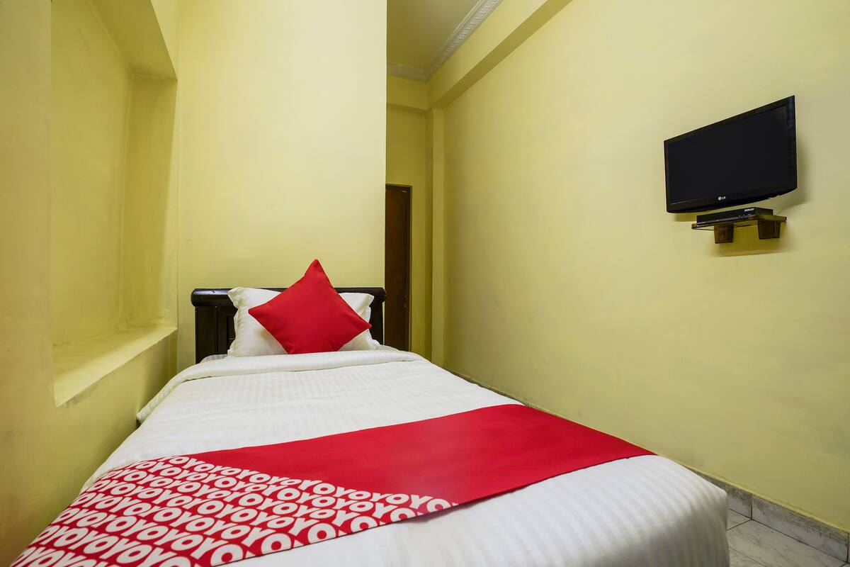A narrow hotel room with a bed, a red pillow, a TV mounted on the wall, and a patterned throw blanket.
