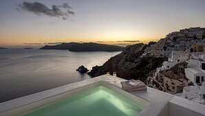A sunset view from a cliff overlooking a coastal town with white buildings, a swimming pool, and two glasses of white wine. - Big Blue Villa by Caldera Houses (Santorini)