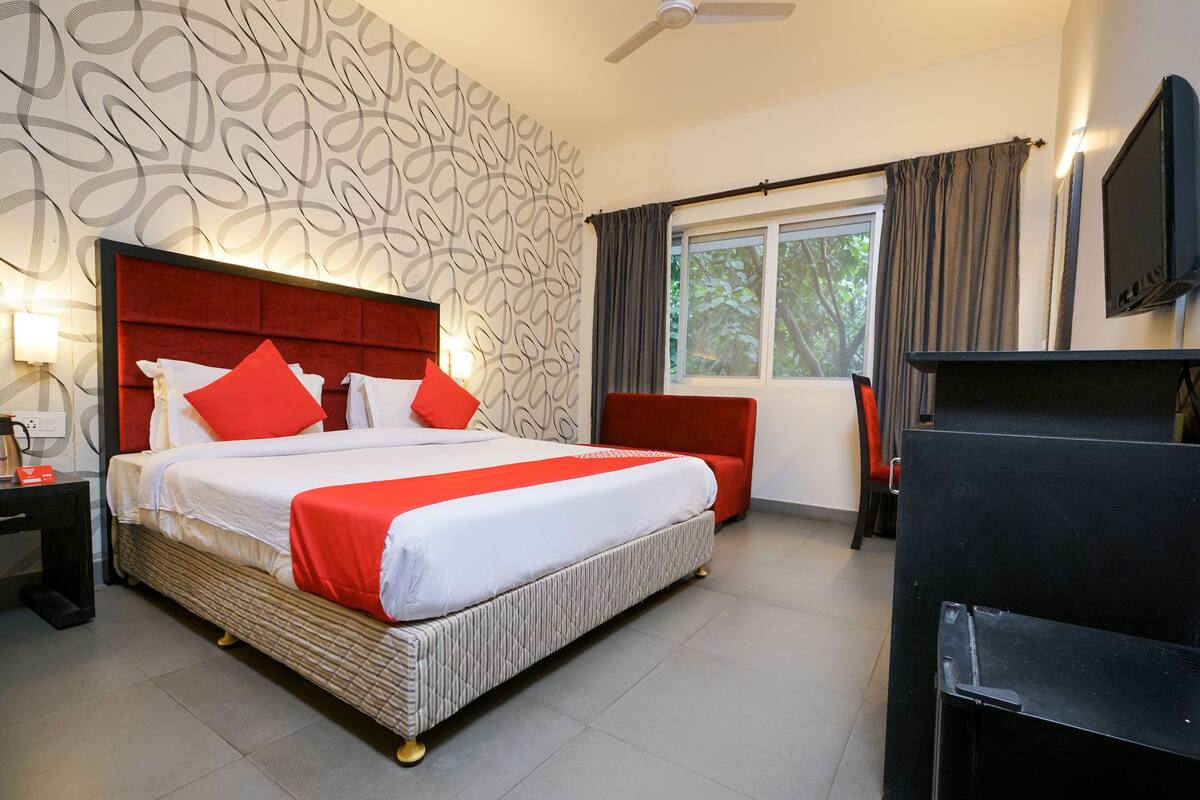 A hotel room with a large bed, a red headboard, a television, and a window with curtains.