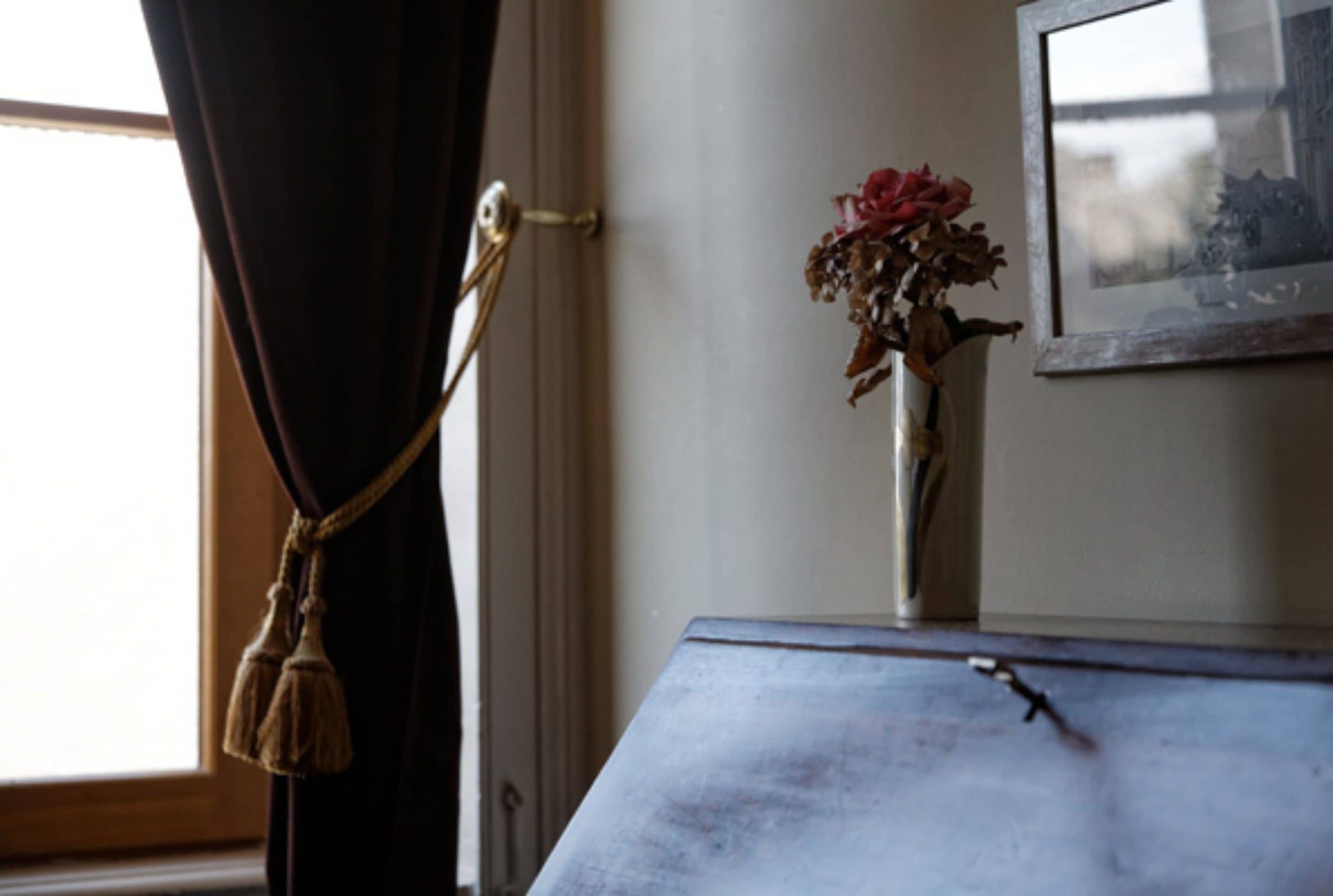 A room with a window, a curtain, a vase with dried flowers, and a framed picture on the wall.