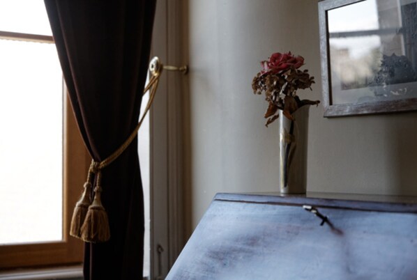 Una habitación con ventana, cortina, un jarrón con flores secas y un cuadro en la pared.