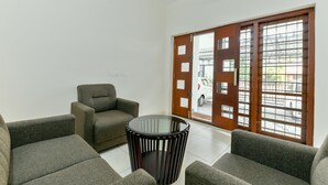 A living room with a round glass table, two sofas, and a wooden door with a view of the street. - OYO 18602 Home Spacious Stay Near Lulu Mall (Kanayannur)
