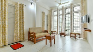 A living room with a sofa, coffee table, and two chairs. There are large windows with curtains, a ceiling fan, and a flat-screen TV mounted on the wall. - OYO 18602 Home Spacious Stay Near Lulu Mall (Kanayannur)