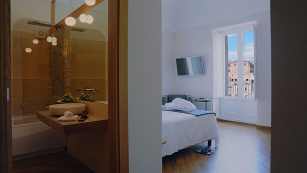 Een moderne badkamer met een grote spiegel, een wastafel en een bad. In de aangrenzende kamer staat een bed met een televisie.